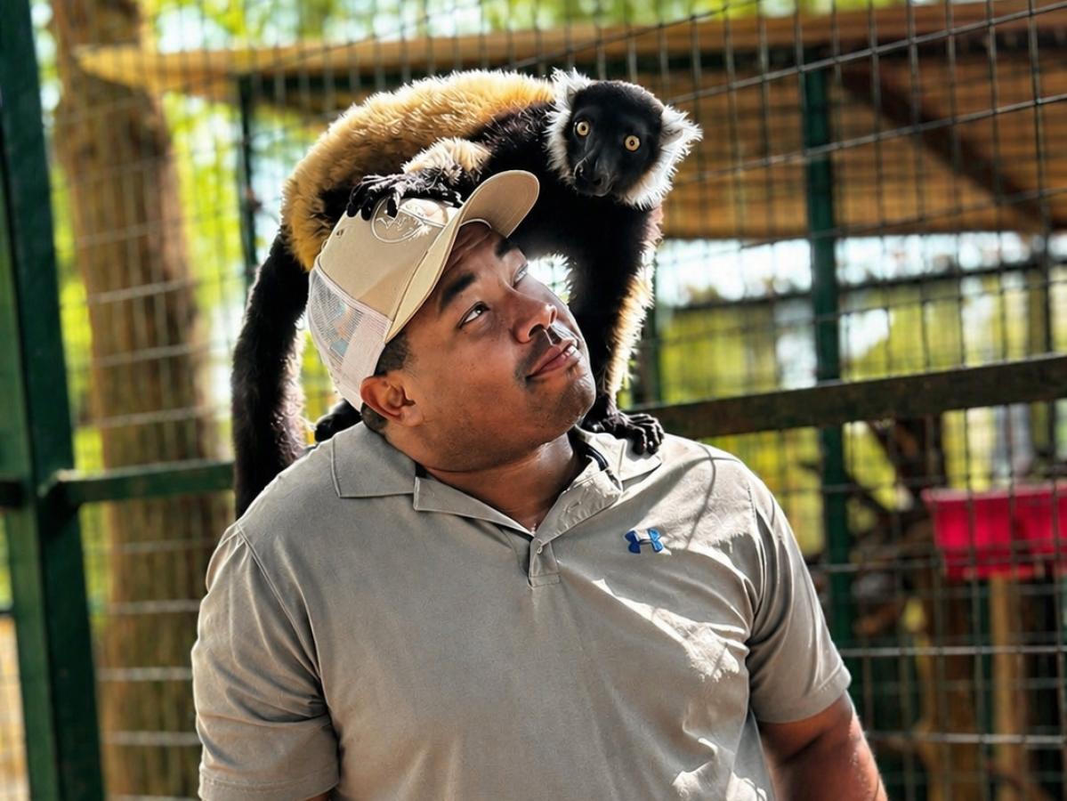 Man with cap looking up at lemur sitting on his shoulder, outdoors.