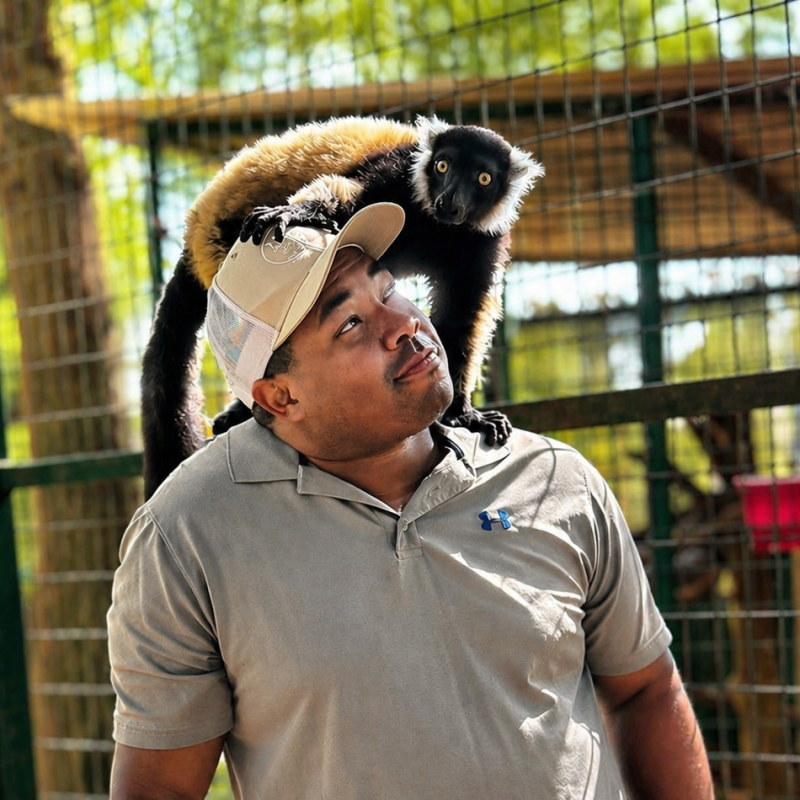 Man with cap looking up at lemur sitting on his shoulder, outdoors.
