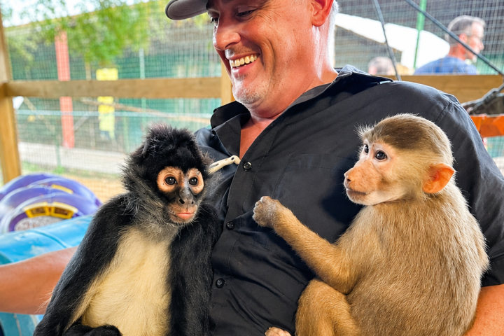 Man smiling with two monkeys sitting on his lap at a zoo enclosure.