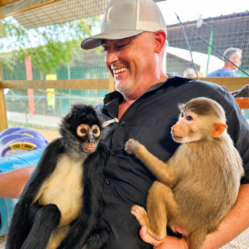 Man smiling with two monkeys sitting on his lap at a zoo enclosure.