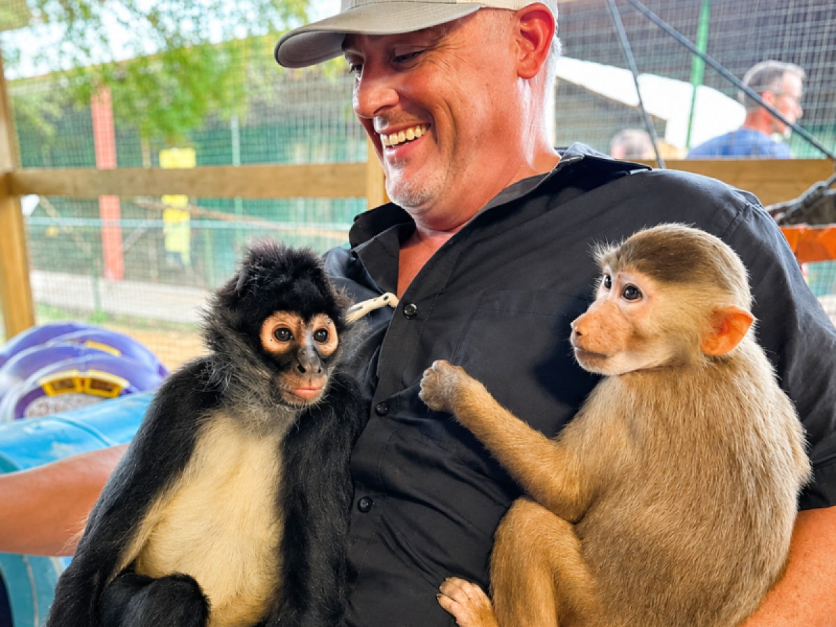 Man smiling with two monkeys sitting on his lap at a zoo enclosure.