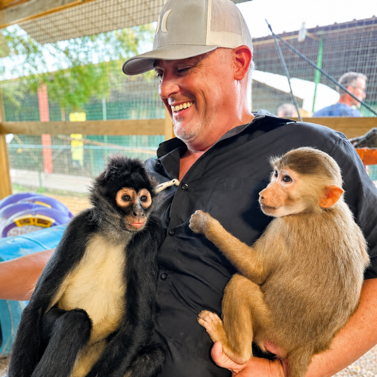 Man smiling with two monkeys sitting on his lap at a zoo enclosure.