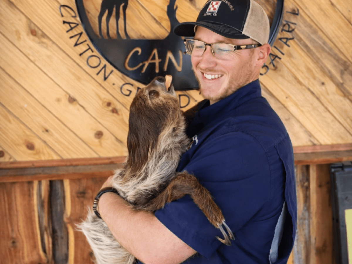 Person in blue shirt holds a sloth in front of a wooden sign.