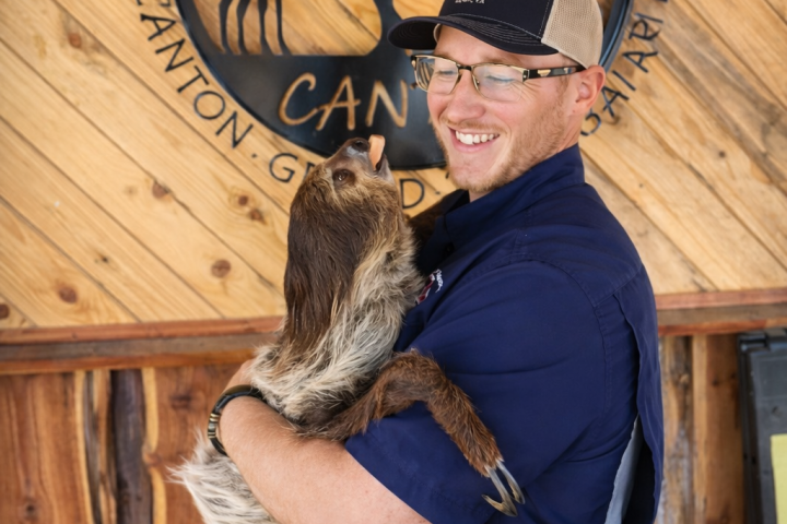 Person in blue shirt holds a sloth in front of a wooden sign.