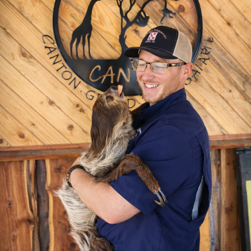 Person in blue shirt holds a sloth in front of a wooden sign.