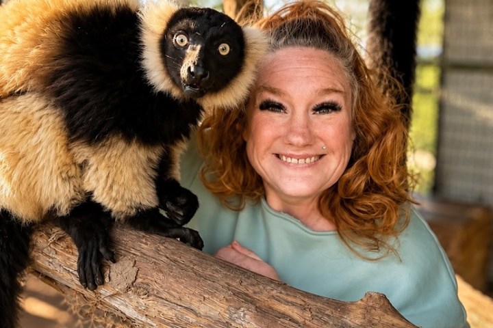 Person smiling beside a black and white lemur on a wooden branch.