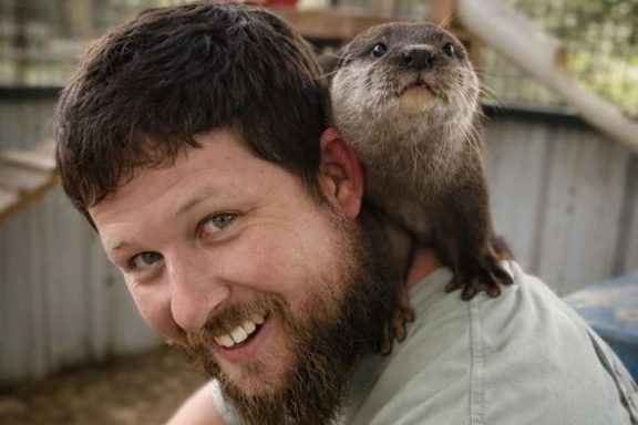 Man smiling with an otter on his shoulder in an outdoor enclosure.