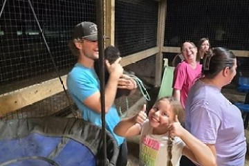 Group of people in an indoor enclosure with a sloth and swinging chair, girl showing thumbs up.