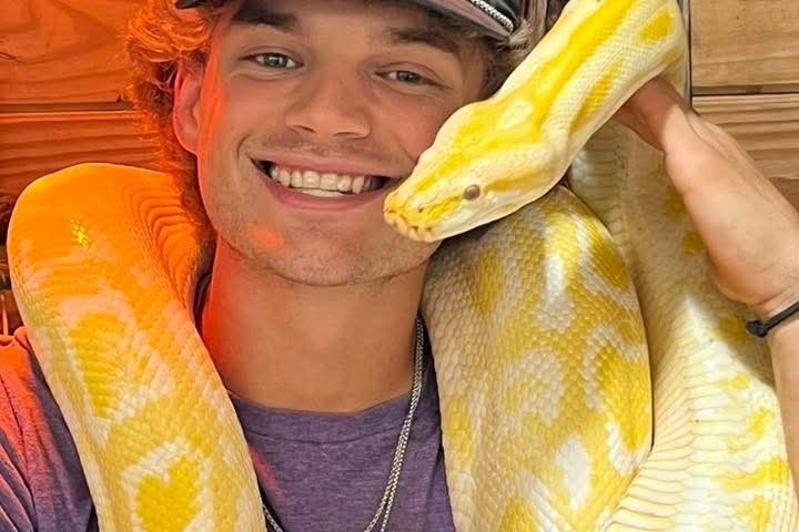 Person smiling, wearing cap, holds large yellow snake around neck.